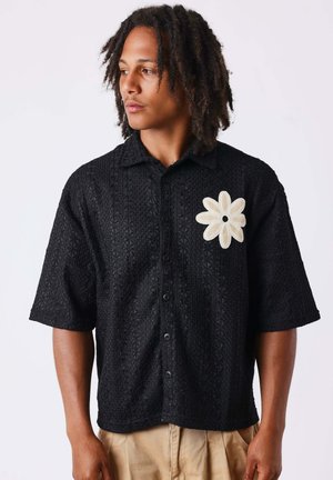 Jeune homme avec des dreadlocks mi-longs portant une chemise noire texturée à manches courtes avec un grand patch fleur blanche sur la poitrine.
