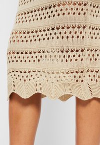 Beiges Strickkleid mit offenen Mustern, gewelltem Saum und abwechselnden horizontalen Streifen aus festen und ausgeschnittenen Abschnitten.