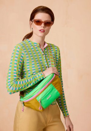 Groene en gele schoudertas met een roze rits, gemaakt van duurzaam materiaal en met een voorvak, gedragen over een patroontrui.