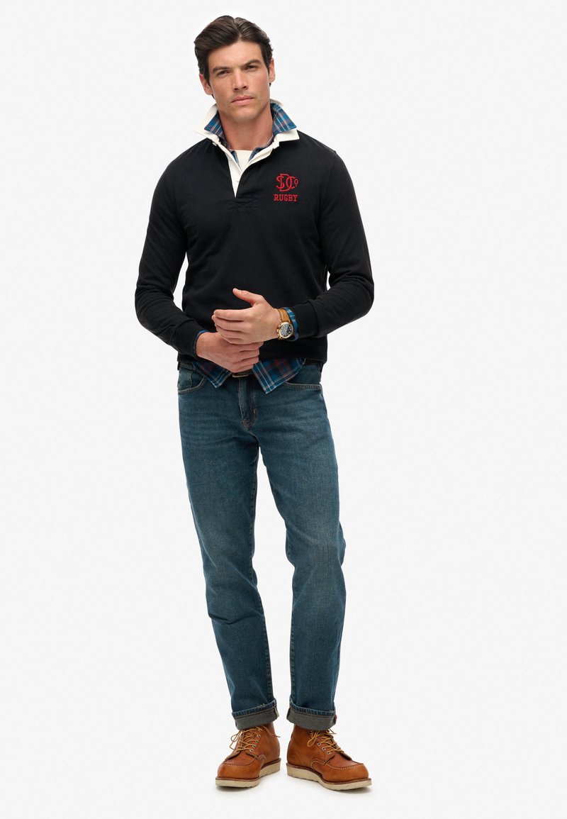 Man in een zwart rugbyshirt over een blauw geruit shirt, blauwe jeans met omslagen en bruine veterschoenen, staand met gekruiste handen.