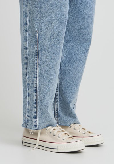 Gai zili džinsi ar brīvu piegriezumu, ar sānu šķeltnēm un kontrastējošām baltām šuvēm, apvienoti ar bēšiem kanvas apaviem.