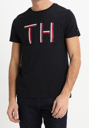 Mann trägt ein schwarzes kurzärmeliges T-Shirt mit rot, weiß und blau gestreiften "TH"-Buchstaben auf der Vorderseite und dunklen Hosen.