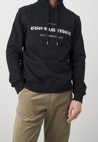 Zwarte hoodie met wit tekstontwerp aan de voorkant, capuchon met trekkoord, geribbelde boorden en zoom, gecombineerd met beige ribfluwelen broek.