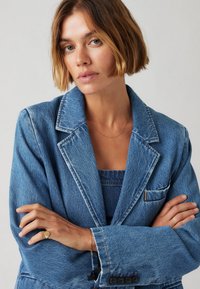 Denim sakO v svetlo modri barvi z odrezanimi klapami, sprednjim žepom in vidnimi šivi. Mehka tekstura, gumbni zaponki na manšetah.