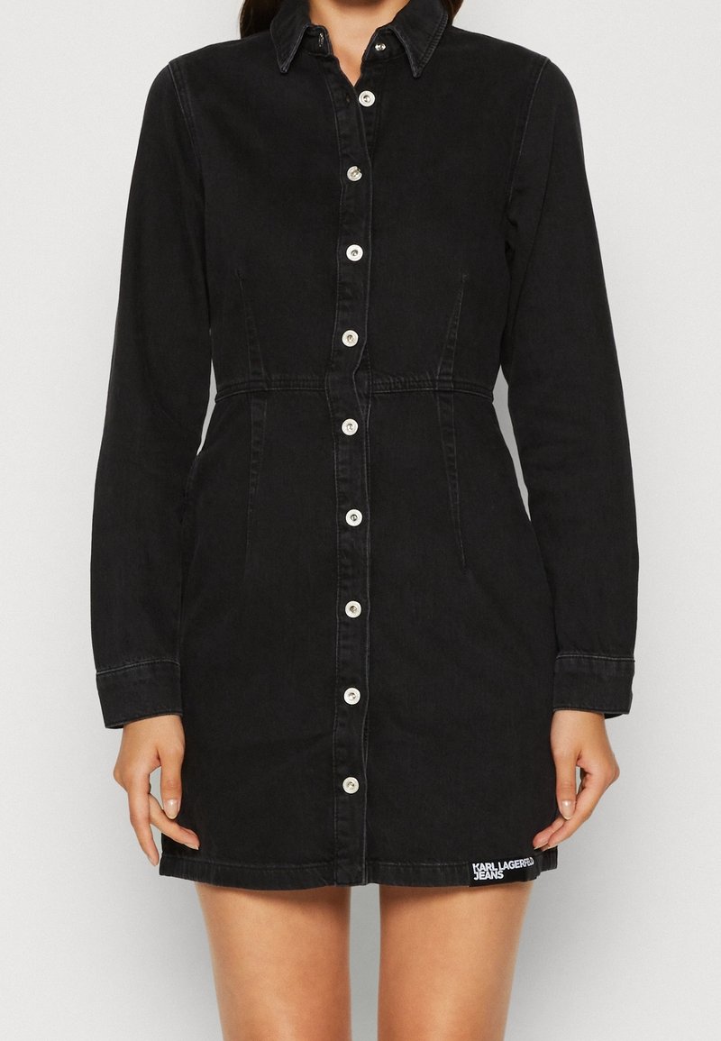 Černé džínové šaty hemžící se dlouhými rukávy, zapínání na knoflíky, špičatý límeček a boční kapsy. Obsahuje bílý logo detail na spodním lemu.