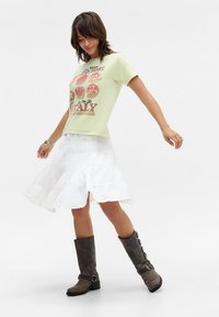Light green graphic t-shirt with circular patterns and text, paired with a white tiered skirt. Brown knee-high boots with side buckles.