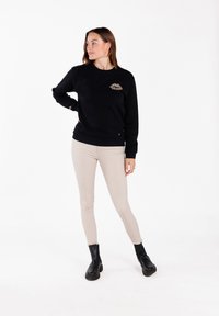 Sudadera negra con cuello redondo y un acento de estampado de leopardo en el pecho, combinada con pantalones skinny beige y botas negras hasta el tobillo.