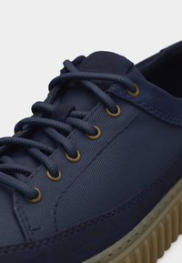 Σκούρο μπλε αθλητικό παπούτσι με υφασμάτινη υφή, στρογγυλές μπρονζέ ρέλια, χοντρές κορδέλες, λεπτομέρειες από σουέτ και ανάγλυφη μπεζ σόλα.