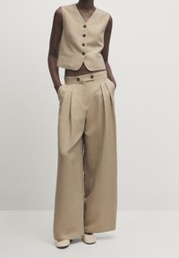 Modella che indossa un gilet beige senza maniche con bottoni e pantaloni a gamba larga a vita alta con tasche laterali, abbinati a scarpe beige, in piedi su uno sfondo neutro.