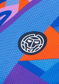 Textil colorido con un patrón de malla que presenta triángulos en azul, púrpura y naranja. Incluye un logo circular negro con un diseño estilizado de cara.