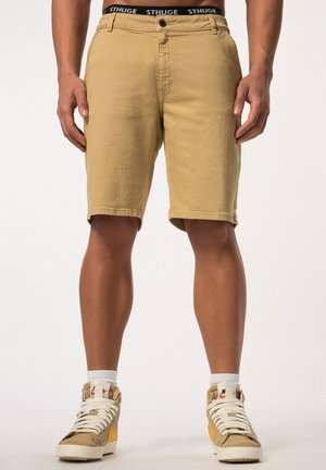 Shorts en kaki en coton avec une texture lisse, dotés d'une fermeture à bouton et d'ourlets roulés. Portés avec des baskets à tige haute.