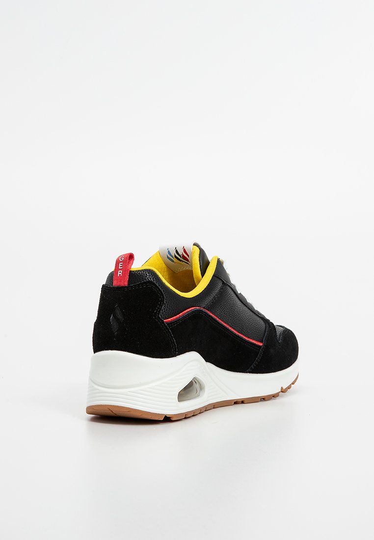 Zwarte suède en mesh sneaker met gele accenten, rode branding op de hak en een witte zool met een zichtbare luchtunit.