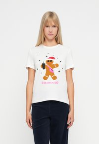 Γυναίκα που φοράει ένα λευκό t-shirt με γραφικό ενός μπισκότου τζίντζερ που κρατάει ένα ποτό, φοράει γυαλιά ηλίου και χριστουγεννιάτικο καπέλο, με το κείμενο "Φαίνομαι Σα να Είμαι Σνακ."