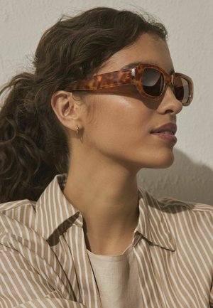 Sieviete ar cirtainiem matiem sasietiem atpakaļ, valkājot bruņurupuča bruņu krāsas saulesbrilles, mazu zelta riņķa auskaru un bēša krāsas svītrainu krekli pret gaišu sienu.