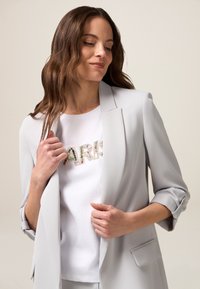 Light gray blazer with rolled sleeves over a white t-shirt featuring “PARIS” in multicolored rhinestones. Smooth fabric texture.