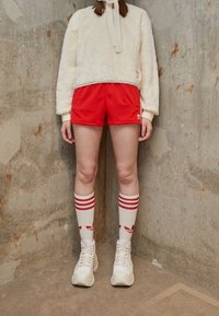 Krämfärgad fuskpäls sweatshirt, röda sportshorts, knästrumpor med röda ränder och logotyper, tjocka vita sneakers. Texturerad, avslappnad stil.