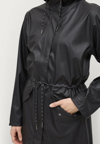 Cappotto impermeabile nero con vita regolabile, davanti con zip, due tasche frontali e cordini testurizzati per il cappuccio. Finitura liscia.