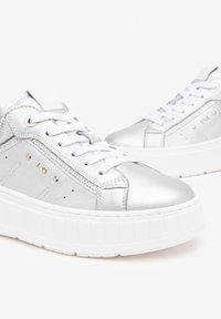 Baskets métalliques argentées avec semelles blanches, présentant un design à lacets, des accents perforés et un minimum de branding sur le côté.