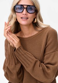 Bruine gebreide trui met geribbelde mouwen, gecombineerd met oversized schildpadbril. Zacht, gestructureerd materiaal, met een relaxed-fit ontwerp.