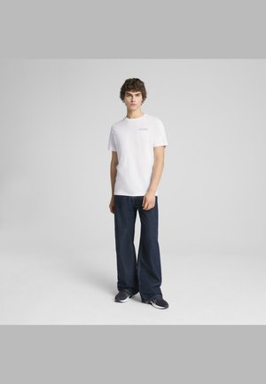 T-shirt bianco in cotone con colletto rotondo e piccolo logo stampato, abbinato a jeans in denim a gamba larga di colore blu scuro e sneakers scure.