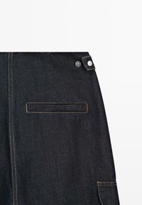 Section de jupe en denim foncé avec poche arrière cousue, patte latérale à bouton-pression et couture verticale visible sur fond blanc.