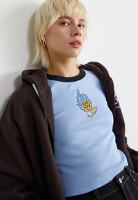 Maglietta fitted blu chiaro con colletto a coste nero, decorata con una grafica di rosa arancione con fiamme blu. Indossata sotto una felpa con zip marrone scuro.