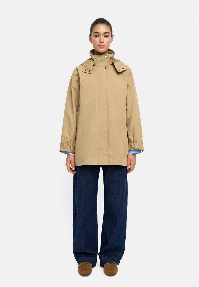 Beżowa, wodoodporna kurtka z wysokim kołnierzem, z zapięciami na napy i elastycznymi mankietami. Stylizowana z luźnymi niebieskimi dżinsami i brązowym obuwiem.