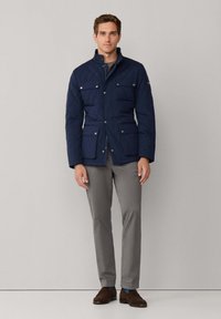 Marineblå kviltet jakke med trykknap- og lynlåslukning, to forlommer, stående krave, båret over en grå skjorte og lysegrå bukser.