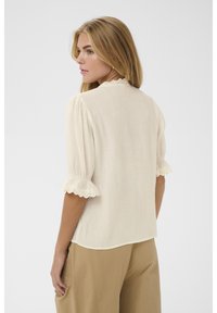 Blondide juustega naine, kes kannab kreemikat tekstuuriga pluusi lamedate ääretega ja heledaid pruune pükse, seisab seljaga vastu, kuid vaatab õla üle.