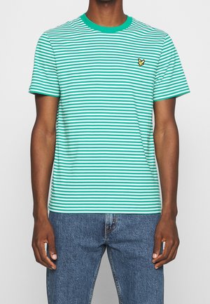 Mann trägt ein kurzärmeliges, grün-weiß horizontal gestreiftes T-Shirt mit einem kleinen gelben Vogel-Logo auf der linken Brust und blaue Jeans.