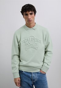 Sudadera verde menta con cuello y logotipo en relieve "SCALPERS", que presenta un diseño de calavera y tibias cruzadas; combinada con vaqueros azules.