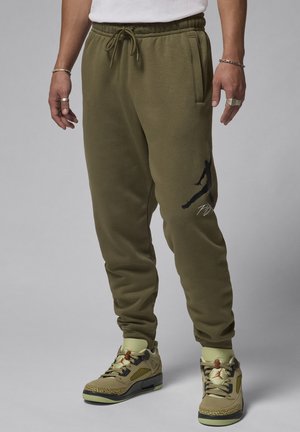 Olive green sweatpants with elastic waistband, side pockets, and black graphic detail. Paired with tan and black patterned sneakers.