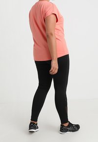 Koral kortærmet top parret med sorte leggings med netpaneler. Sportssko fuldender outfittet og fremhæver en strømlinet silhuet.