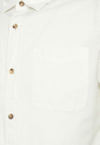 Camicia bianca con bottoni, realizzata in tessuto leggero. Presenta una tasca sul petto, colletto arrotondato e bottoni in legno naturale. Design minimalista.