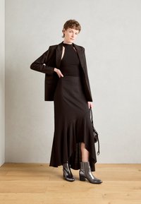 Μαύρη εφαρμοστή σαλοπέτα πάνω από ένα μαύρο αμάνικο τοπ και μια μακριά, φούστα με φούσκωμα. Μαύρες μπότες έως τον αστράγαλο με χοντρό τακούνι. Λείο ύφασμα και καθαρές γραμμές.