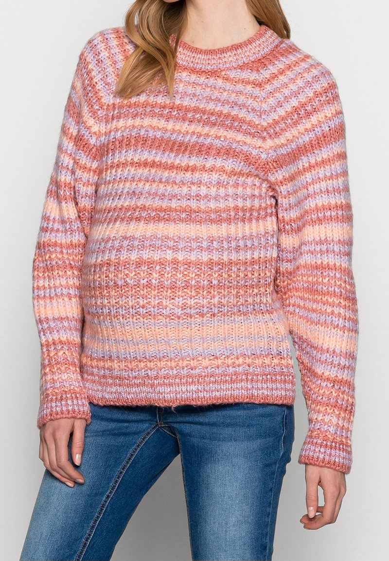 Femme portant un pull en maille côtelée à rayures horizontales dans des tons de rose, pêche et lavande, associé à un jean bleu.