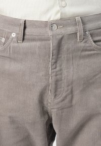 Brune corduroy bukser med lodrette striber, med en metal knap, bæltestropper og forlommer. Stoffet ser blødt og tekstureret ud.