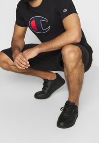T-shirt in cotone nero con grande logo rosso e bianco, abbinato a pantaloni corti neri e sneakers nere. La texture appare liscia e informale.