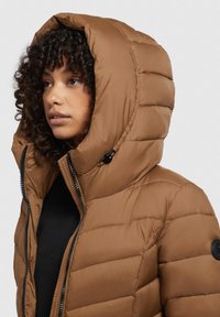 Hnědá vyp padded bunda s vysokým límcem a velkou kapucí. Obsahuje černý zip a kulatou nášivku s logem na levém rukávu.