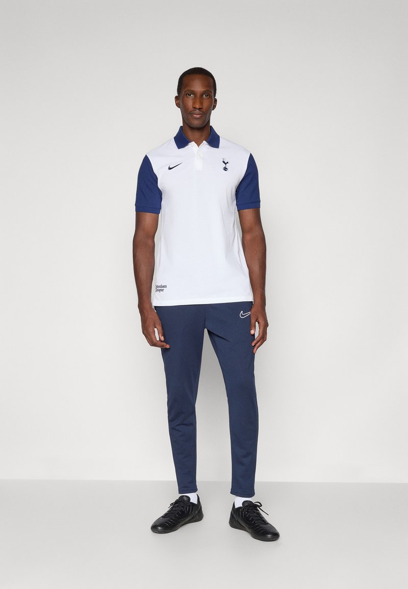 Polo shirt blanc avec manches marines, ornée d'un logo sportif et de boutons. Associée à un pantalon de jogging marine et des chaussures de sport noires.