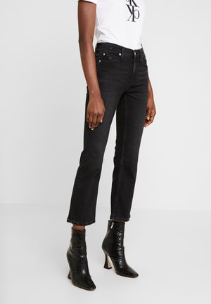 Personne portant un jean noir droit, des bottines noires à talons avec une texture crocodile et une chemise blanche avec un logo noir.