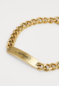 Kultaketju, jossa on suuria lenkkejä, ja joka sisältää sileän, tasaisen nimilaatan kaiverrettuna "Ed Hardy". Kiiltävä metallipinta ja tukevat rakenteet.