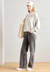 Grijze sweatshirt met roze logo, zwarte wijde jeans en witte sneakers. Accessoires zijn een beige tote bag en een witte baseballcap.