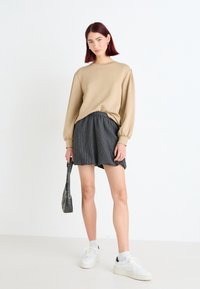 Beige Übergröße Pullover mit Ballonärmeln, kombiniert mit dunklen gestreiften Shorts und weißen Sneakern. Das Model hält eine kleine schwarze Tasche.