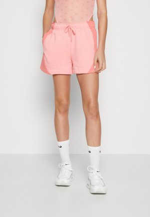 Personne portant un short de sport rose avec cordon de serrage, des chaussettes blanches montantes et des baskets blanches Nike, debout devant un fond blanc uni.