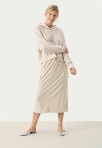Felpa beige con cappuccio e tasca frontale, abbinata a una lunga gonna beige con tasche laterali. Il modello indossa ballerine argentate con punta.