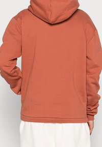 Mikina v rezavé barvě, vyrobená z měkké tkaniny, s prostornou kapucí a volným střihem. Dlouhé rukávy a ribbované manžety dotvářejí design.