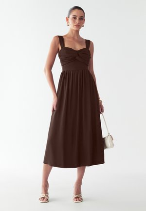 Hnedé midi šaty s širokými ramienkami a skrúteným živôtikom, splývavá sukňa. Modelka nosí sandále na remienky a drží malú kabelku. Hladká tkanina.