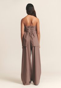 Hnědý top bez ramínek s nařaseným detailem na zádech, doplněný kalhotami s širokými nohavicemi, oba z jemné texturované látky. Minimalistický design s čistými liniemi.