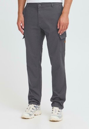 Man wearing dark gray cargo pants with flap pockets and white athletic shoes standing against a plain background.